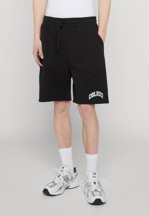 Persona che indossa pantaloncini neri con coulisse con il logo "Colucci" sulla gamba sinistra, calzini bianchi e scarpe da ginnastica bianche New Balance, in piedi davanti a uno sfondo semplice.