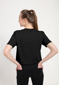 Černé bavlněné tričko s krátkými rukávy, kulatým výstřihem a volným střihem. Nosené s přiléhavými černými kalhotami. Hladká textura, minimální design.