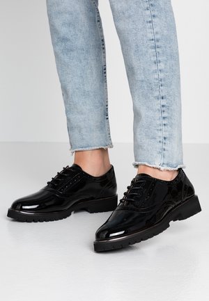 Schwarze Brogue-Schuhe aus Lackleder mit dicken Gummisohlen, getragen zu hellblauen, kurz geschnittenen Jeans.