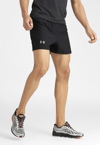 Mies, joka on pukeutunut mustiin Under Armour -shortseihin ja juoksukenkiin, seisoo valkoista taustaa vasten yhdellä jalalla hieman nostettuna.