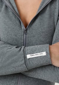 Persona con un suéter gris de cremallera y los brazos cruzados, mostrando una etiqueta blanca en el puño que dice "COOL Cozy Comfy."