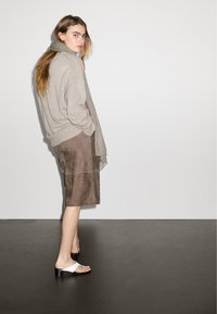 Sweter w kolorze taupe, luźny krój, zestawiony z brązową zamszową spódnicą do kolan i białymi oraz czarnymi sandałami. Gładka tekstura, prosty design.