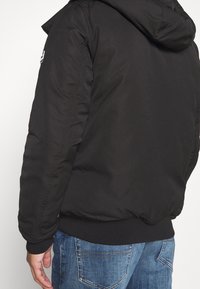 Person som bär en svart hujacka med ribbade muddar och midjeband, kombinerad med blåa jeans i denim, sedd bakifrån och från sidan.