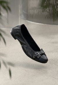 Chaussure plate en cuir noir avec un bout arrondi, dotée d'une sangle ornée de œillets en argent et d'un détail de boucle. Texture lisse et design minimaliste.