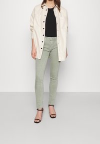 Camisa chaqueta larga de crema con botones negros, combinada con jeans skinny verde claro y sandalias de tiras negras. Tela suave, corte relajado.