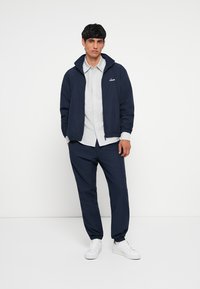 Jūrininkų spalvos zip-up striukė su apykakle, po ja baltos dryžuotos marškiniai ir atitinkamos jūrininkų spalvos kelnes. Apranga turi švarią, minimalistinę dizaino estetiką.