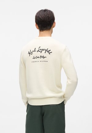 Krém pulóver bordázott szegéllyel és mandzsettával, a hátoldalán fekete hímzett szöveggel: "Karl Lagerfeld," "La Côte D'Azur," és "Francia Riviéra."
