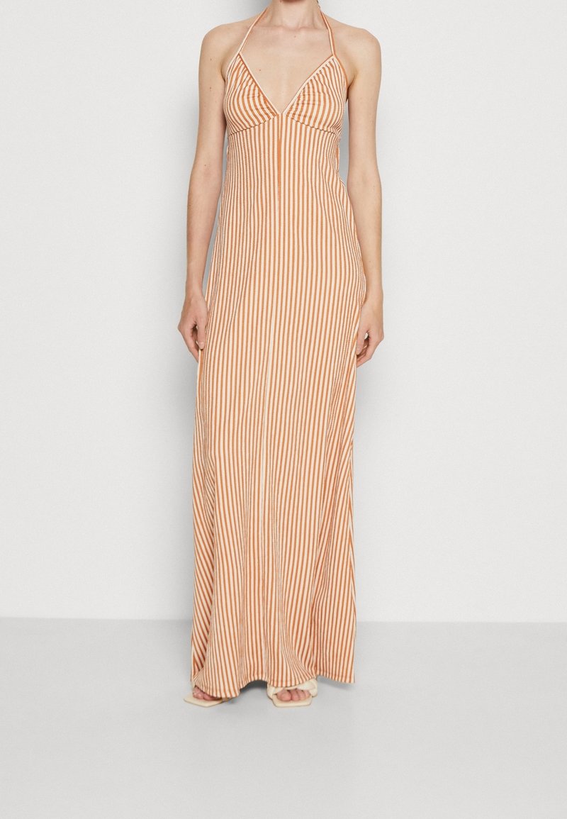 Femme portant une robe longue au sol à rayures verticales orange et blanches, avec un dos nu et un décolleté en V profond, et des sandales.