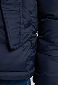 Jachetă puffer de culoare bleumarin, cu textură matlasată, având un mic etichetă neagră cu un "P" portocalie pe partea stângă, aproape de cusătură.