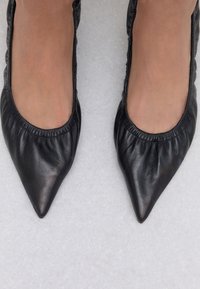 Black leather flats with a pointed toe, featuring gathered detailing around the opening. Smooth texture and minimalistic design.