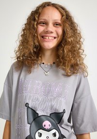 Szara, dużych rozmiarów koszulka z nadrukiem dużego bohatera kreskówkowego oraz fioletowym tekstem. Projekt zawiera błyszczące akcenty i miękką teksturę materiału.