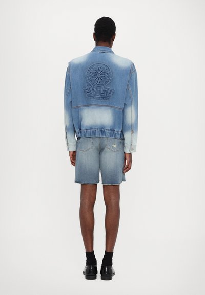 Τζάκετ από μπλε τζιν με ξεθωριασμένη υφή, με λογότυπο στο πίσω μέρος. Συνδυασμένο με ανοιχτόχρωμα τζιν σορτς. Ορατά μαύρα παπούτσια και κάλτσες.