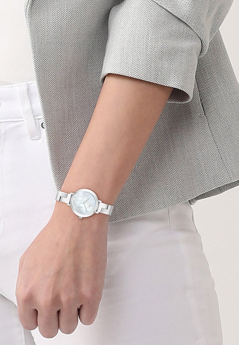 Mujer con un reloj pulsera de metal plateado con esfera clara, manga de chaqueta texturizada gris y pantalones blancos.
