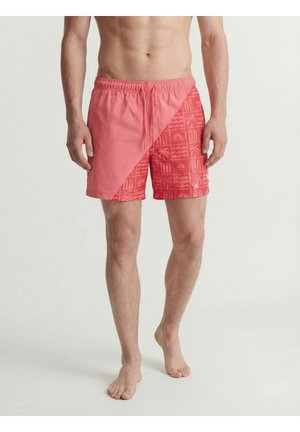 Muž nosi koralové plavecké šortky s diagonálnym rozdeleným vzorom, naľavo jednofarebné, napravo tropický vzor, stojí bosý na jednofarebnom pozadí.