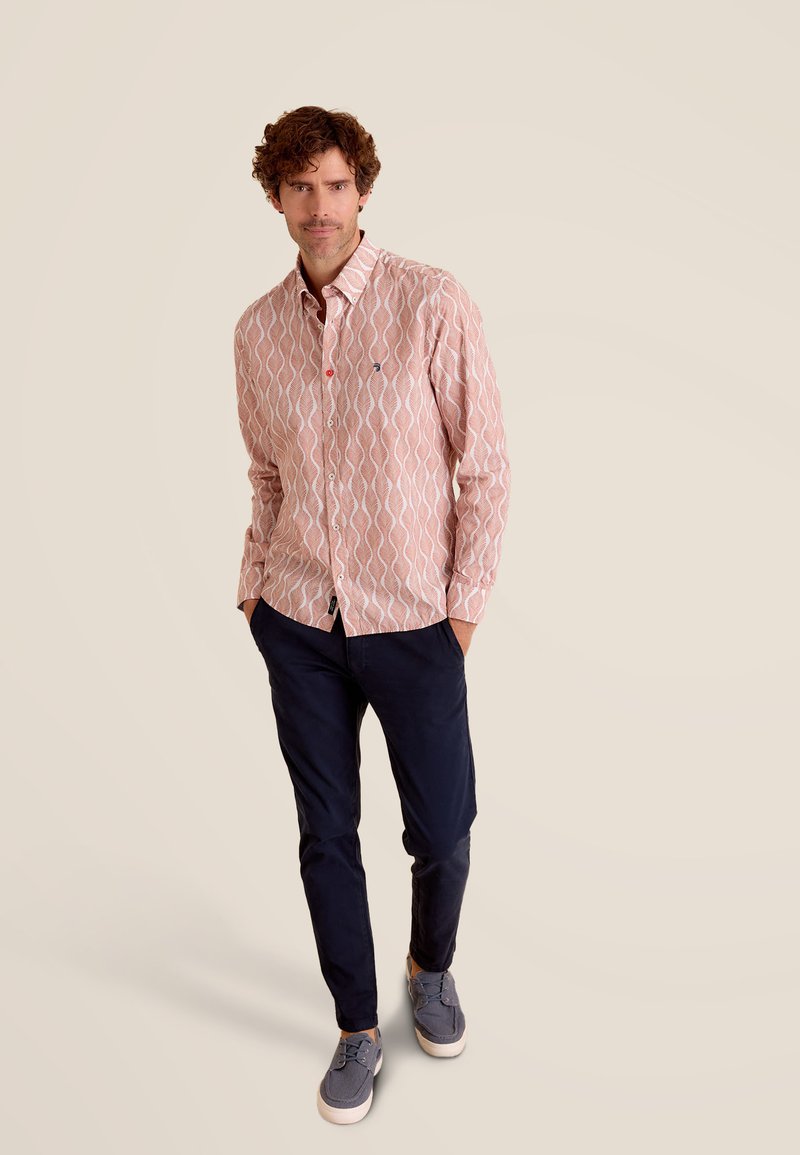 Langarmshirt in lichtroze met een witte golfpatroon, met knoopsluiting en een rode accentknop. Gecombineerd met donkerblauwe broek en grijze schoenen.