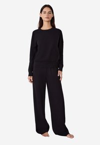 Černý pletený svetr s kulatým výstřihem a dlouhými rukávy, kombinovaný s volnějšími černými kalhotami s širokými nohavicemi a minimalistickým designem.