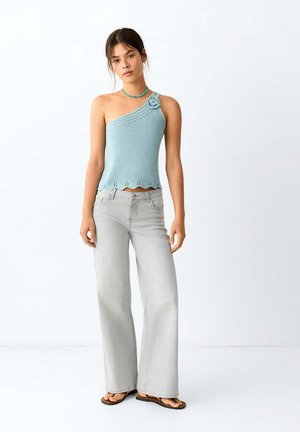 Junge Frau trägt ein hellblaues, einseitiges Stricktop mit Blumendetail, hellgraue, weit geschnittene Jeans und schwarze Flip-Flops und steht still.
