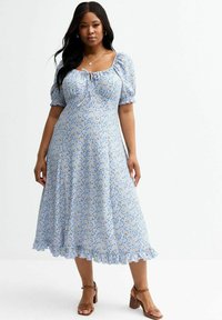 Robe midi bleue à fleurs avec manches bouffantes et décolleté carré. Présente un nœud à l'avant et un ourlet froncé. Portée avec des talons beiges.