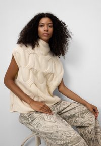 Beżowy, dziany bezrękawnik z golfem o fakturowanym wzorze falującym, zestawiony z marmurkowymi, jasnoszarymi spodniami w abstrakcyjny wzór.