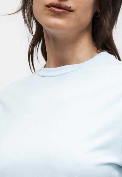 Γυναίκα με καστανά μαλλιά μέχρι τους ώμους, που φοράει ένα γαλάζιο μπλουζάκι με στρογγυλή λαιμόκοψη, με μια ελιά πάνω από το πάνω χείλος.