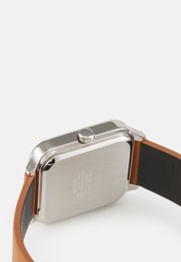 Boîtier de montre en acier inoxydable carré avec une finition polie, bracelet en cuir marron et doublure intérieure noire. Couronne positionnée sur le côté.