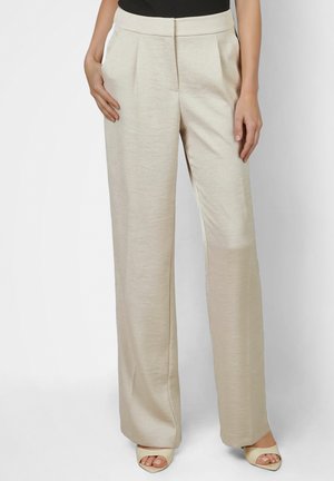 Woman wearing beige high-waist wide-leg trousers with front pleats, side pockets, black top, and beige open-toe heels, standing straight.