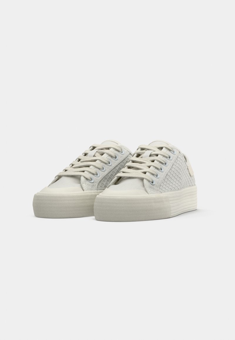 Baskets en toile blanches présentant un côté texturé, des lacets plats, une pointe arrondie et une semelle en caoutchouc épaisse avec un motif rayé.