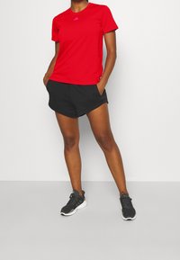 Camiseta atlética roja con mangas cortas y escote redondo, combinada con pantalones cortos negros y zapatillas grises. Logo de adidas en rosa en la camiseta.