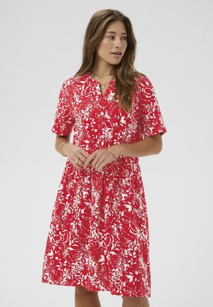 Kobieta w czerwono-białej sukience w kwiaty, sięgającej do kolan, z krótkimi rękawami, stojąca na jednolitym jasno-szarym tle.