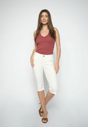 Vrouw staande met één been gekruist, draag een mouwloze roestkleurige top, witte capri-broek en bruine open-toe hakken, tegen een effen witte achtergrond.