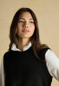 Must must-valge nööpidega särk, mille peal on must kudumivest, millel on detailne äär. Juuksed on tumedad ja lainelised, taust on neutraalne.