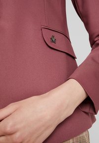 Gros plan d'une manche de veste mauve et d'un rabat de poche avec une petite épingle décorative métallique et une main visible près de la taille.