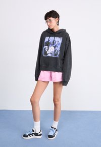 Felpa nera con cappuccio e finitura sbiadita, caratterizzata da una stampa colorata. Abbinata a pantaloni corti rosa e scarpe da ginnastica nere con lacci azzurri.