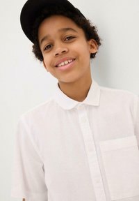 Camicia bianca a maniche corte con colletto, dotata di taschino frontale e tessuto testurizzato. Indossata con un cappellino nero.