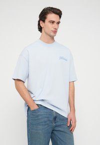 Νεαρός άνδρας με σκουρόχρωμα μαλλιά, που φοράει ανοιχτό μπλε φαρδιά μπλούζα με μικρό λογότυπο στο στήθος και μπλε τζιν, στέκεται με το ένα χέρι στην τσέπη.