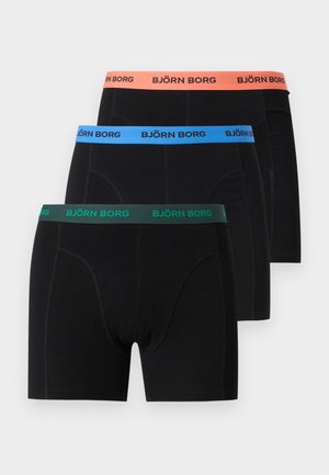 Magas férfi mezítláb áll a szőnyegen, fekete boxernadrágot visel narancssárga derékpánttal, amelyen a "BJÖRN BORG" felirat látható, oldalra néz egy egyszínű háttér előtt.