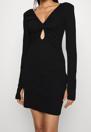 Robe noire à manches longues en tissu côtelé, avec un décolleté en V et une découpe en forme de clé au niveau de la poitrine. Coupe ajustée et longueur au-dessus du genou.
