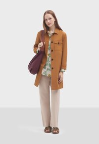Cappotto lungo in cotone marrone con bottoni neri, camicia floreale verde sotto, pantaloni beige e sandali con cinturini marroni. Borsa a spalla viola.