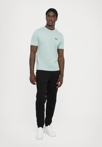 Myntegrønn kortermet T-skjorte med EA7-logo, kombinert med svarte bukser og hvite sneakers. Stoffet ser jevnt ut, med en avslappet passform.
