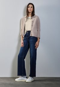 Hellgraue, fuzzy Jacke mit Knöpfen vorne, cremefarbener Pullover, dunkelblaue weit geschnittene Jeans und weiße Turnschuhe an einem Modell vor einem schlichten Hintergrund.