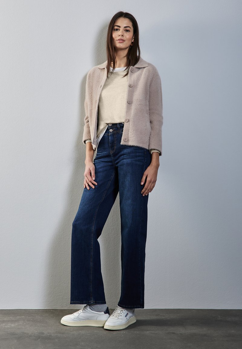 Hellgraue, fuzzy Jacke mit Knöpfen vorne, cremefarbener Pullover, dunkelblaue weit geschnittene Jeans und weiße Turnschuhe an einem Modell vor einem schlichten Hintergrund.