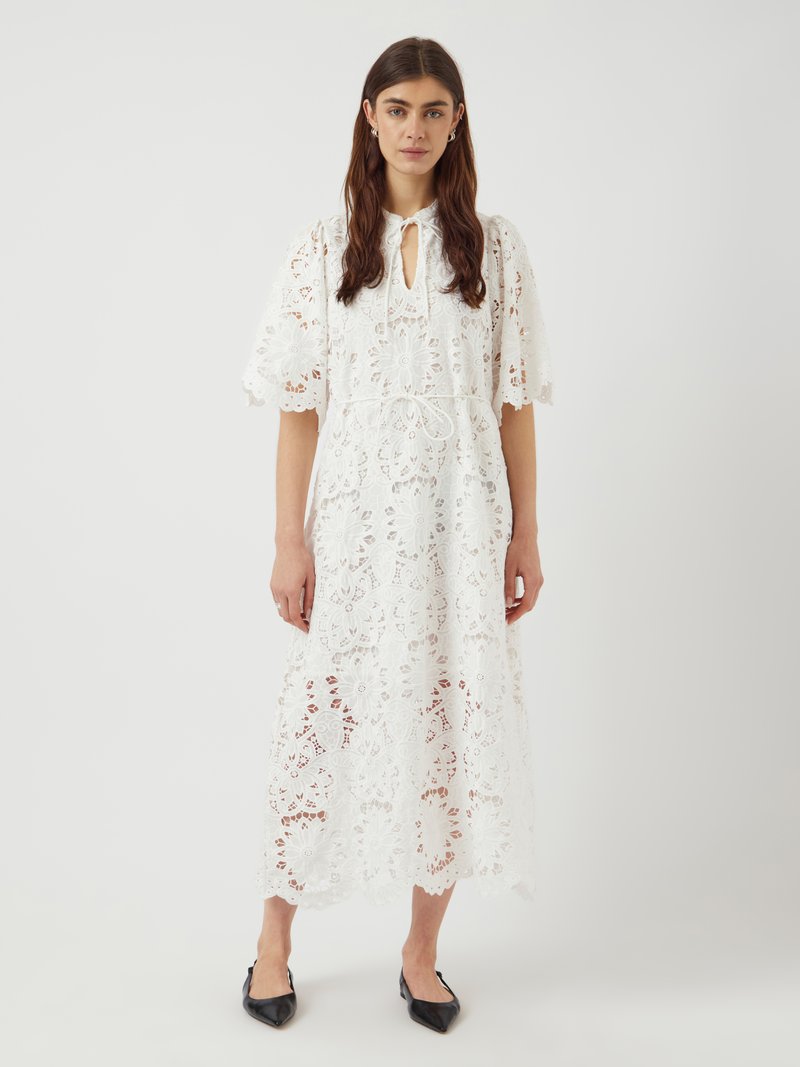 Λευκό δαντελένιο φόρεμα με λουλουδάτο σχέδιο, κοντά φουσκωτά μανίκια και κλείσιμο με κορδόνι στο λαιμό. Μέσου μήκους με προπέτασμα στο τελείωμα. Συνδυασμένο με μαύρα παπούτσια.
