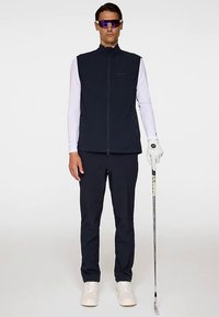 Marinblå ärmlös golfväst över en vit långärmad skjorta; matchad med marinblå byxor och vita skor. Handskar på vänster hand, håller i en golfklubba.