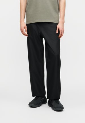 Inimene, kes kannab lahtiseid musti pükse, musti libisevaid kingi ja oliivrohelist t-särki, seisab ühevärvilise heleda tausta ees.