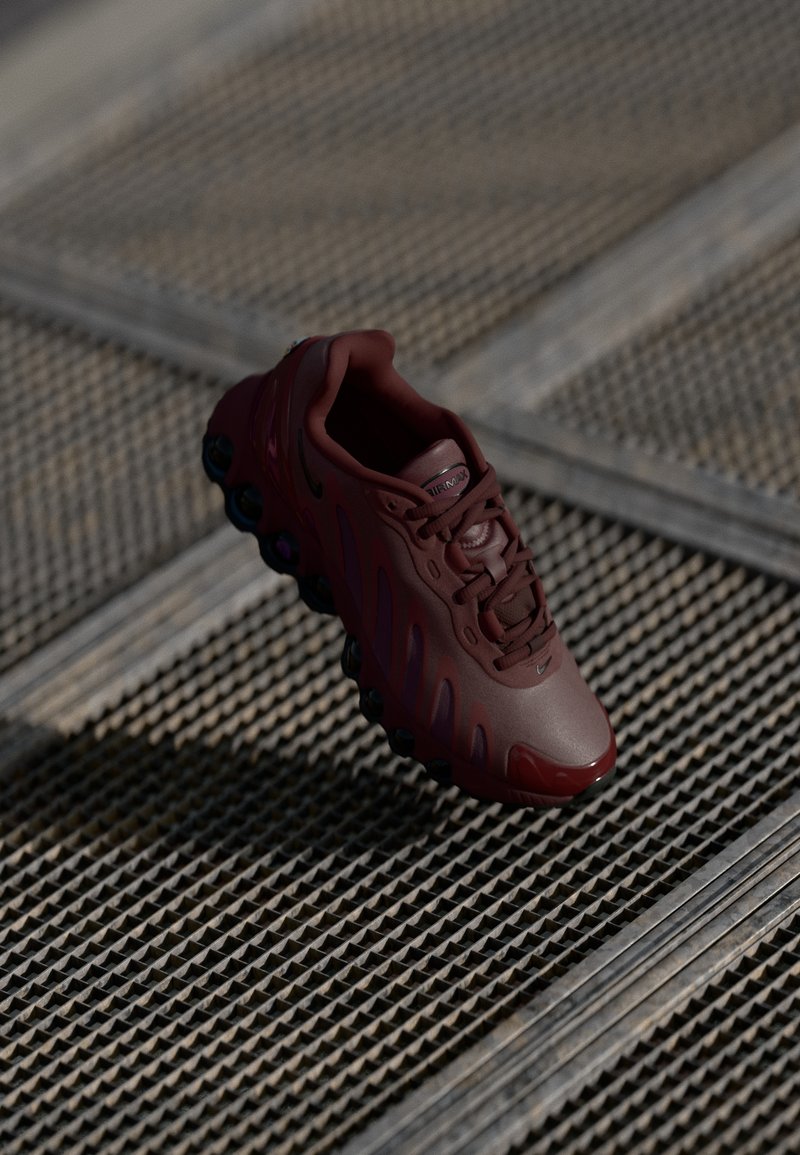 Bordowe buty sportowe z teksturowaną cholewką, wyraźnymi sznurówkami i widocznymi poduszkami powietrznymi. Charakteryzują się smukłymi liniami i błyszczącą osłoną na palcach.