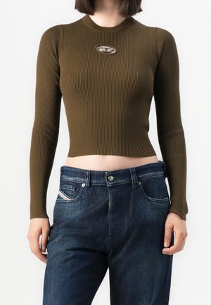 Femme portant un haut court en maille côtelée vert olive à manches longues avec une découpe métallique ovale et un jean taille haute bleu foncé.