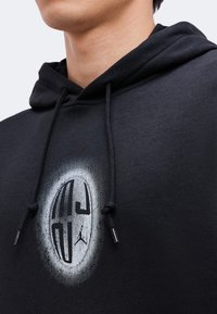 Personne portant un sweat à capuche noir avec un motif circulaire blanc et des cordons noirs, visible du cou jusqu'au milieu de la poitrine.