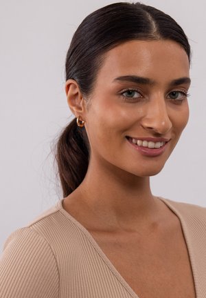 Bronzen creolen, geribbelde beige top, gladde huid, lichtbruin haar in een paardenstaart, neutrale achtergrond. Groene ogen, subtiele glimlach.