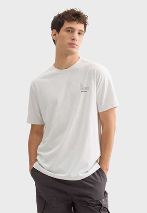 Camiseta blanca de mangas cortas con un corte relajado, que presenta un pequeño logo gris en el lado izquierdo del pecho. Tejido suave y textura lisa.
