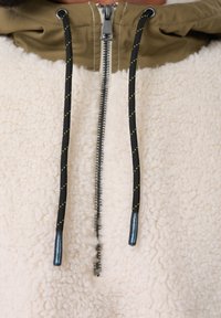 Fliso megztinis su kreminės spalvos tekstūra, alyvuogių žalumos gaubtu, juodais raišteliais su geltonais akcentais ir sidabrine užtrauktuko uždanga.